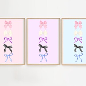 Op de afbeelding: Drie ingelijste kunstdrukken met aquarel strikken in roze, wit, paars, zwart en blauw. Elke print heeft een andere pastel achtergrond: roze, lila en lichtblauw. De strikken zijn verticaal gerangschikt.