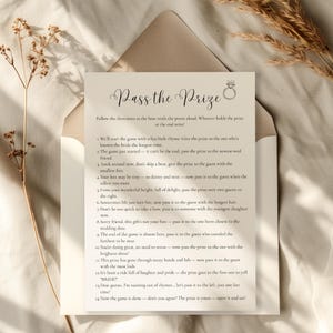 May include: A cream-colored card with the text "Pass the Prize" in elegant script, featuring a ring graphic. The card is inside a matching envelope, with a list of instructions for a bridal shower game. Dried flowers are in the background.
