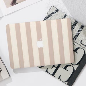 Op de afbeelding: Een harde laptophoes met een verticaal streeppatroon in afwisselende tinten beige en crème. De hoes heeft een centrale uitsparing die het zilveren Apple-logo op het deksel van de laptop onthult en het ontwerp over het gehele oppervlak bedekt.