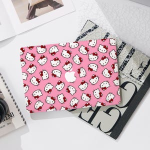 May include: A pink laptop cover featuring a repeating pattern of Hello Kitty faces with red bows. The cover has a white Apple logo in the center. The design is set against a white background with other decorative items.