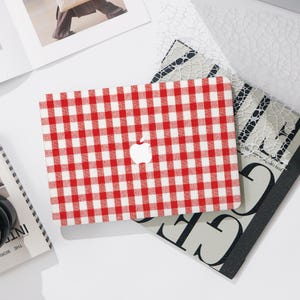 Op de afbeelding: Een rood-witte geruite laptophoes met een wit Apple-logo. De hoes ligt op een stapel boeken en decoratief papier. De hoes heeft een klassiek picknickkleedpatroon.