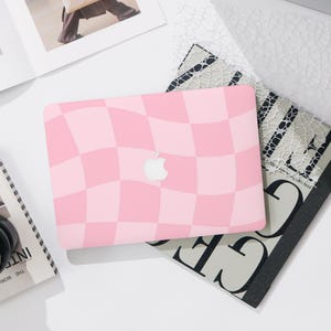 May include: Pink and light pink checkered laptop case with the Apple logo. The case is on top of a magazine and other items on a white surface. The case has a wavy checkerboard pattern.