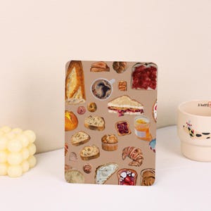 May include: A rectangular, tan-colored notebook cover featuring a watercolor illustration of various baked goods, including bread, croissants, muffins, and sandwiches. A cup of coffee and a jar of jam are also depicted. A cream-colored mug is in the background.