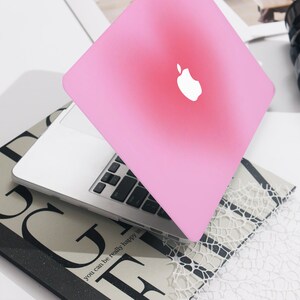 May include: A pink laptop with a white Apple logo sits open on a book. The laptop is a light pink colour. The book has the words "you can be really happy in" printed on it.