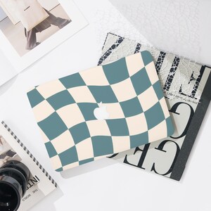 May include: A laptop case with a teal and cream checkered pattern. The Apple logo is visible in the center. The case is on top of a stack of books and magazines. The background is white.