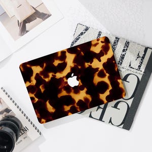 May include: A laptop computer with a tortoiseshell pattern cover. The cover is a mix of dark brown and tan colors. The Apple logo is visible on the cover. The laptop is on top of a stack of magazines and other items.