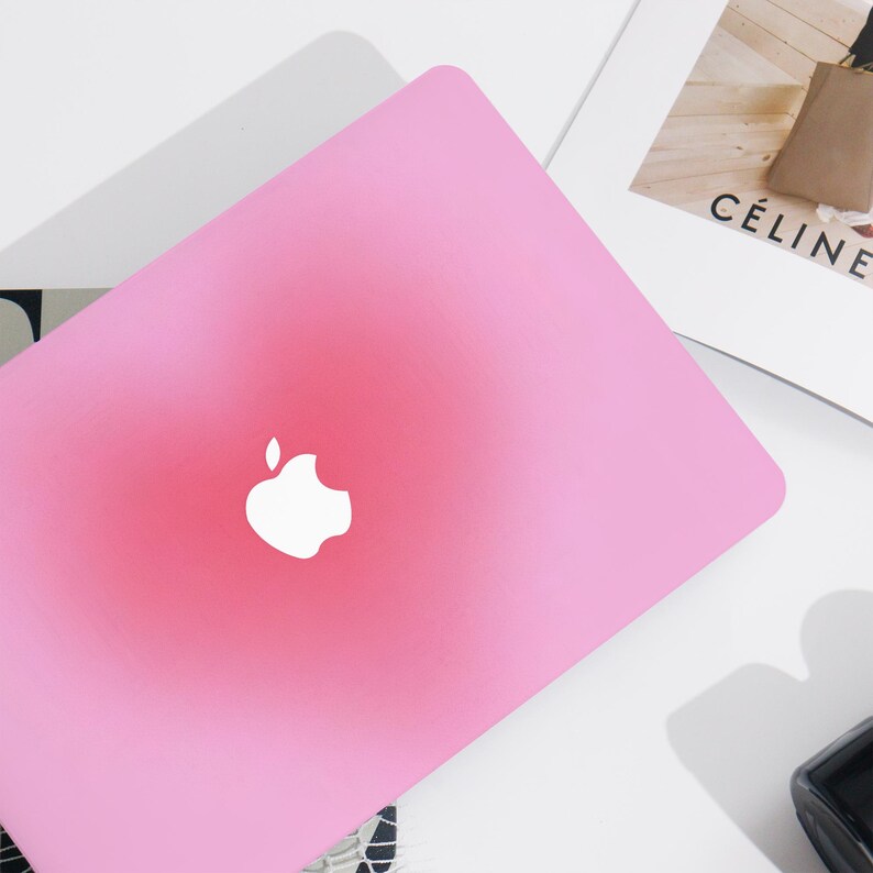May include: A pink laptop featuring the Apple logo in white. The laptop is set against a white backdrop, with a magazine and a black object visible. The magazine displays the word "C&Eacute;LINE".