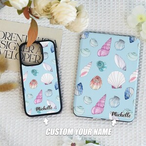 Può includere: Una custodia per telefono e una custodia per tablet azzurro chiaro con un motivo a conchiglie nei toni del rosa, bianco e blu. Le custodie hanno un bordo nero e il nome "Michelle" è stampato su di esse. È visibile anche la frase "CUSTOM YOUR NAME".