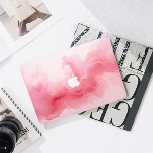 May include: A pink laptop case with a swirling, abstract design. The case features a white Apple logo and is surrounded by books and other stationery items on a white surface.