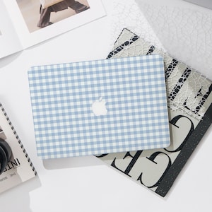May include: A laptop case with a light blue and white gingham pattern, featuring the Apple logo. The case is placed on top of books and magazines. The background is white.