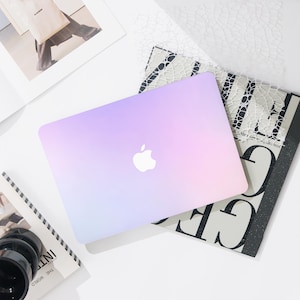 Könnte beinhalten: Ein Laptop mit Farbverlauf in Lila und Rosa mit dem Apple-Logo auf dem Deckel. Der Laptop liegt auf einem Stapel Bücher und Zeitschriften. Der Hintergrund ist weiß.