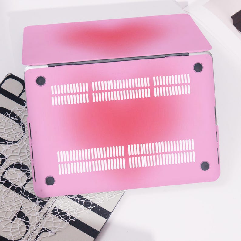 May include: A pink laptop case with a gradient design, shifting from a darker red shade in the middle to a lighter pink at the borders. It has ventilation slots and black rubber feet.