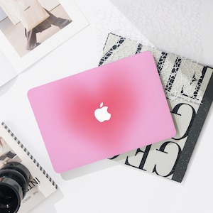 May include: A pink laptop case with a gradient design, featuring the Apple logo in white. The case is on top of a book with black and white text and other stationery items. The overall aesthetic is modern and minimalist.