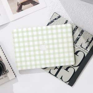 May include: A light green and white gingham pattern laptop case with the Apple logo. The case is on top of a stack of magazines and a decorative paper with a lace design.