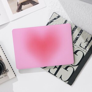 May include: A pink laptop case featuring a heart-shaped design in the centre. The case is placed on a stack of books and magazines. The heart design is a darker shade of pink.