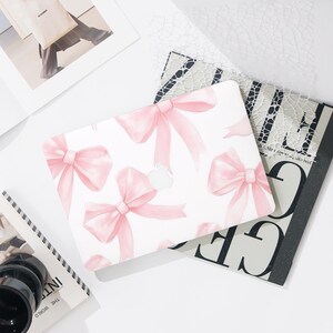 Puede incluir: Funda blanca para portátil con un estampado de lazos rosas en acuarela. La funda tiene un recorte para el logotipo de Apple. Se ven otros artículos de papelería, como una revista y un cuaderno.