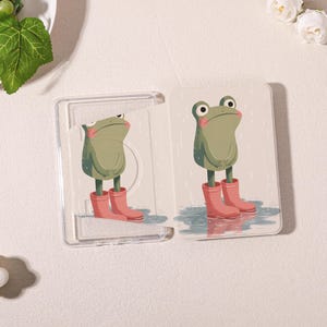 May include: A clear plastic card holder with a whimsical design featuring a cartoon frog wearing pink rain boots. The frog is depicted in a rainy scene, adding a playful touch. The card holder is open, showcasing the frog illustration.