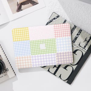 May include: A pastel-colored laptop case with a patchwork design. The case features squares of yellow, pink, blue, green, and purple gingham patterns. A white Apple logo is centered on the case. The case is on a white surface with other items.