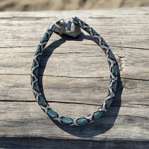 May include: A beaded necklace with a geometric pattern in teal, black, and silver. The necklace is displayed on a weathered wooden surface, with a blurred sandy background.