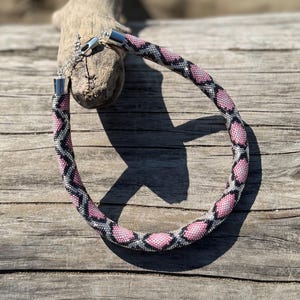Op de afbeelding: Een kralenketting met een slangachtig patroon. De ketting heeft een herhalend ontwerp van roze, zwarte en zilveren kralen. De ketting wordt getoond op een stuk drijfhout tegen een houten achtergrond.