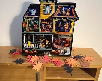 May include: A wooden haunted house decoration with multiple windows and doors, each depicting a different spooky scene. The house is painted yellow with black trim and sits on a wooden table with fall leaves scattered around it.