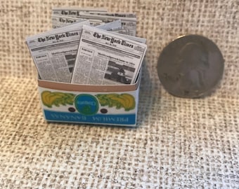 May include: A miniature cardboard box with a blue and white label that reads "PREMIUM BANANAS" and a banana graphic. The box is filled with miniature newspapers that read "The New York Times".