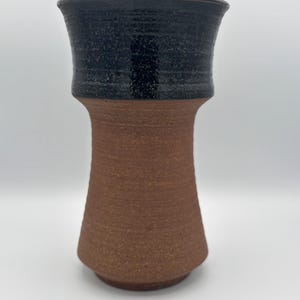 May include: A handmade ceramic vase with a two-tone design. The top section is a dark, speckled black, while the lower portion is a textured, reddish-brown. The vase has a flared top and a cylindrical body, perfect for displaying flowers or as a decorative piece.