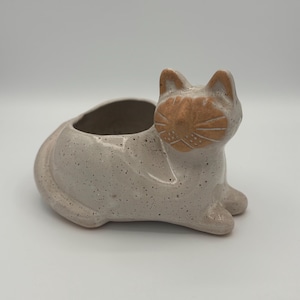May include: A ceramic cat-shaped planter in a speckled off-white color. The cat has a tan face with etched details and a small opening for planting. The planter is designed to sit on a flat surface.