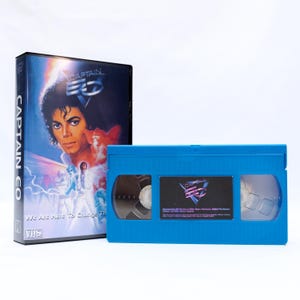 Puede incluir: Una cinta VHS azul y su estuche para "Captain EO" con Michael Jackson. El estuche tiene una imagen colorida de Jackson y el título. La cinta es azul brillante con una etiqueta.