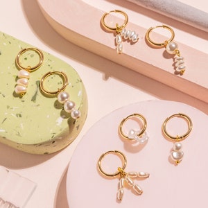 May include: Six pairs of gold hoop earrings with pearl drop charms. The earrings are displayed on pink and green surfaces.