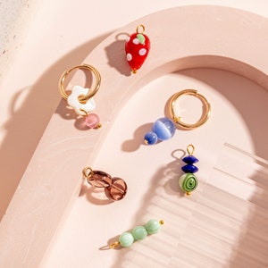May include: A set of six gold hoop earrings with various charms, including a strawberry, a flower, a blue and white swirl, a green and blue swirl, a brown bead, and a green bead.