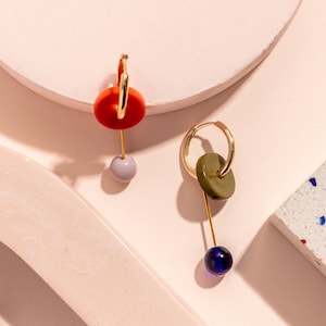 Puede incluir: Un par de pendientes de aro dorados con formas geométricas colgantes. Un pendiente tiene un círculo rojo y una esfera de color morado claro, mientras que el otro tiene un círculo verde y una esfera de color azul oscuro.