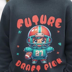 Puede incluir: Una sudadera negra con una ilustración de dibujos animados de un jugador de fútbol americano. El jugador lleva un casco y sostiene un balón. El texto "FUTURE DRAFT PICK" está impreso en rojo y naranja. La camiseta del jugador muestra el número 21.
