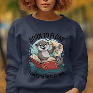 May include: Navy blue sweatshirt featuring a cartoon otter holding a pencil and scroll, sitting on an open book. The text reads "BORN TO FLOAT, FORCED TO GO TO SCHOOL". The design is in shades of blue, brown, and red.