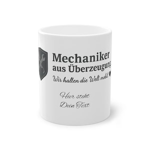 Puede incluir: Taza de cerámica blanca con el texto "Mechaniker aus Überzeugung" y "Wir hallen die Welt mobil" en negro. Un escudo con una llave inglesa está en el lado izquierdo. La parte inferior de la taza tiene el texto "Hier steht Dein Text".