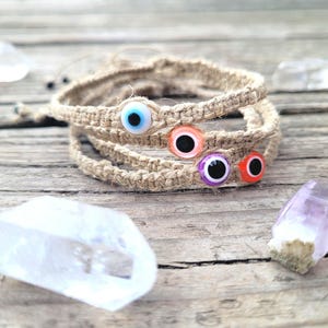 May include: Three woven hemp bracelets, each adorned with a colorful evil eye bead. The beads feature a blue, orange, purple, and red eye design. The bracelets are stacked on a wooden surface, with crystals in the background.