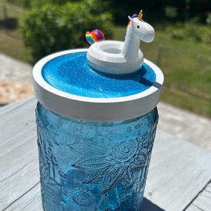 Puede incluir: Un tarro de vidrio azul con una tapa blanca y un unicornio en un flotador arcoíris en una piscina azul de resina. El tarro tiene un diseño floral.