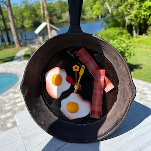 May include: A black cast iron skillet clock with a red and yellow egg design and red bacon strips. The clock hands are red and yellow.