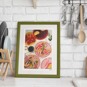 May include: A framed art print featuring a colorful illustration of food, including tacos and other dishes. The artwork is set against a white brick wall, with a wooden dish rack and kitchen utensils visible in the background. The frame is green.