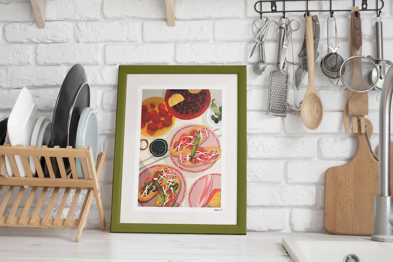 May include: A framed art print featuring a colorful illustration of food, including tacos and other dishes. The artwork is set against a white brick wall, with a wooden dish rack and kitchen utensils visible in the background. The frame is green.