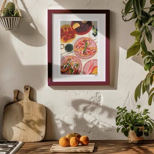 May include: A framed print of a colorful food illustration hangs on a white wall. The artwork depicts various dishes, including plates of food with toppings. A wooden countertop with oranges and cutting boards is in the foreground.