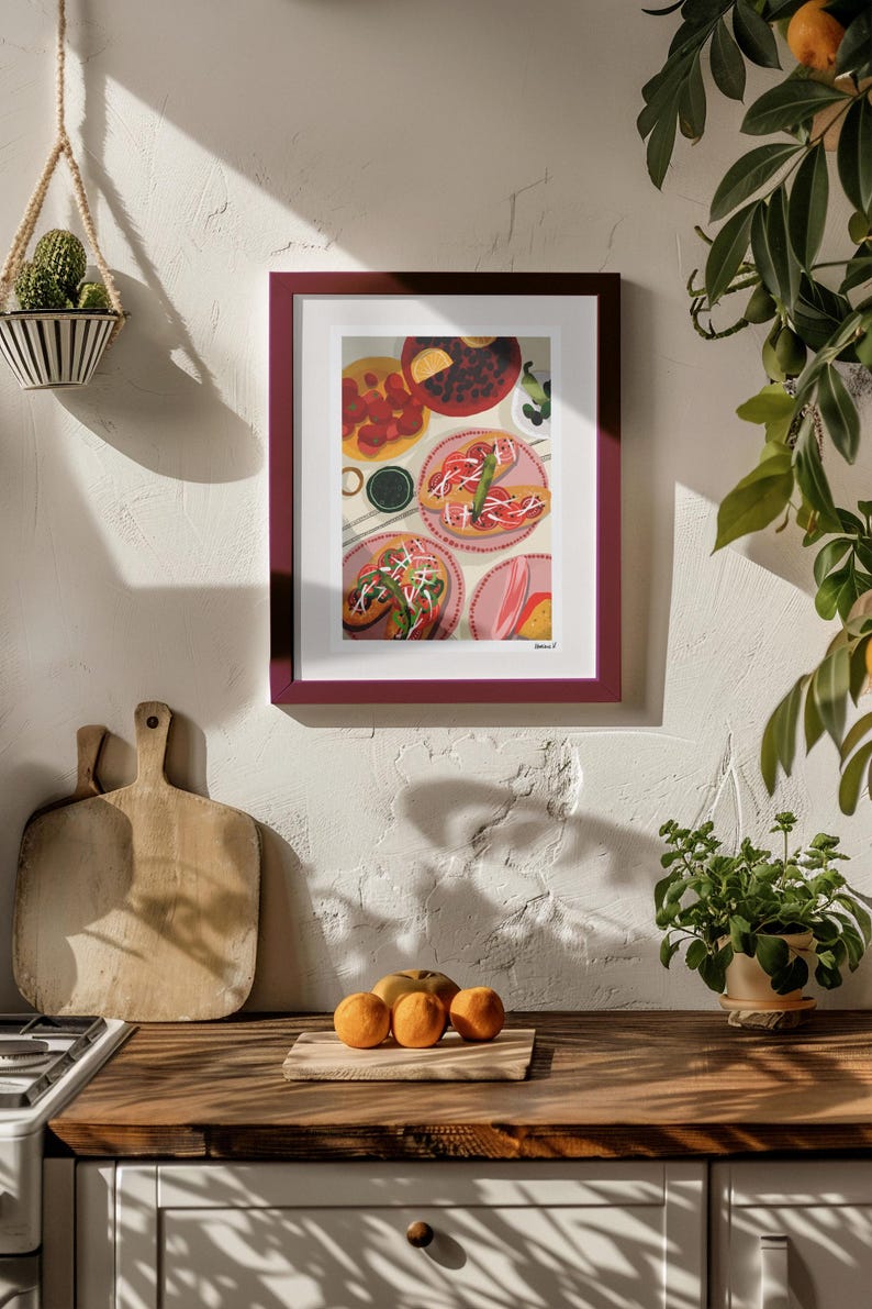 May include: A framed print of a colorful food illustration hangs on a white wall. The artwork depicts various dishes, including plates of food with toppings. A wooden countertop with oranges and cutting boards is in the foreground.