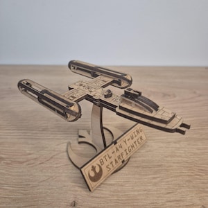 Puede incluir: Maqueta de madera de un caza estelar BTL-A4 Y-wing, con detalles cortados con láser. El modelo se exhibe sobre un soporte de madera con el texto "BTL-A4 Y-WING STARFIGHTER" y un logotipo. El color es marrón claro.