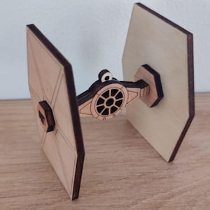 May include: A wooden TIE fighter model, with two large, light-coloured panels and a central cockpit section. The model is made of light-coloured wood with dark brown edges and accents, showcasing a laser-cut design. The model is a miniature.