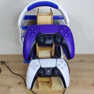 May include: A wooden gaming controller stand holding a purple and a white controller. The stand also holds a white and blue headset. The controllers are stacked on top of each other. The stand is on a wooden surface.