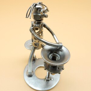 Könnte beinhalten: Eine silberne Metallskulptur einer Person, die Trompete spielt. Die Skulptur besteht aus Muttern, Schrauben und anderen Metallteilen. Die Person steht auf einem runden Sockel.