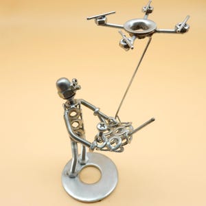 May include: A silver metal sculpture of a person holding a remote control and a drone flying above. The sculpture is made of nuts, bolts, and other metal pieces.