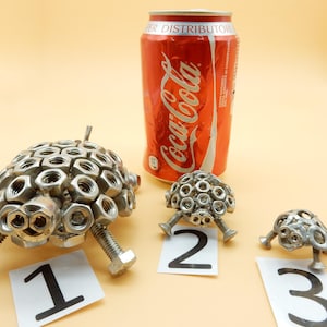 May include: Three metal turtle sculptures made from nuts and bolts are displayed next to a red Coca-Cola can. The turtles are labeled 1, 2, and 3. The can is approximately 4.8 inches tall.