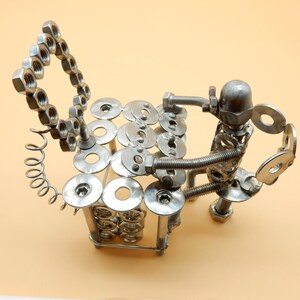 May include: A silver metal sculpture of a person sitting on a chair made of nuts and bolts. The chair has a back made of nuts and a seat made of washers. The person is holding a washer in each hand.