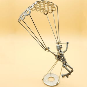 Puede incluir: Una escultura de metal plateado de una persona haciendo paracaidismo con un paracaídas hecho de arandelas de metal. La escultura está sobre una base redonda.